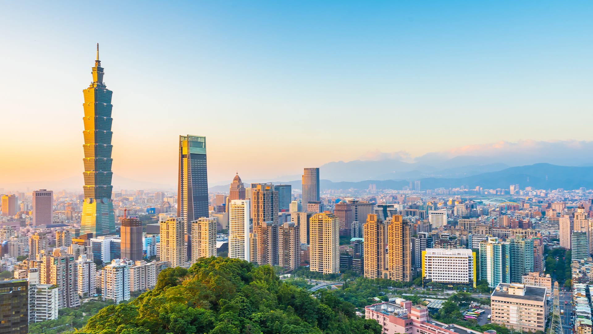 Panoramic view of Taipei city skyline at sunset, featuring the tall Taipei 101 skyscraper surrounded by modern high-rise buildings, with green hills in the foreground and mountains in the background under a clear sky.