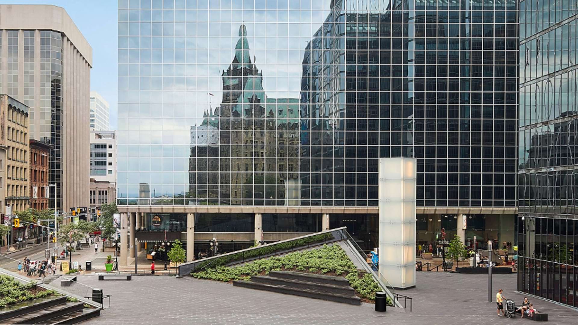A modern urban plaza with dark tiled pavement and landscaped terraces featuring green plants. The plaza is surrounded by tall glass office buildings, one of which reflects an older, castle-like stone building with green roofs. People are walking and sitting around the area, and there are trees and street signs visible along the adjacent road.