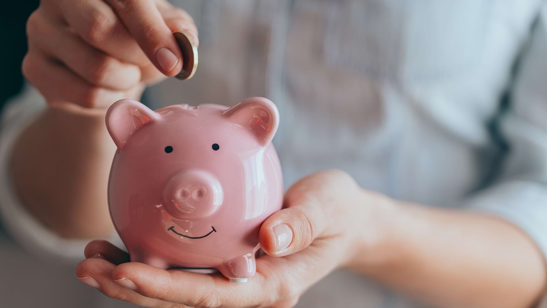 : A close-up of a person's hands putting a coin into the slot of a smiling pink ceramic piggy bank.