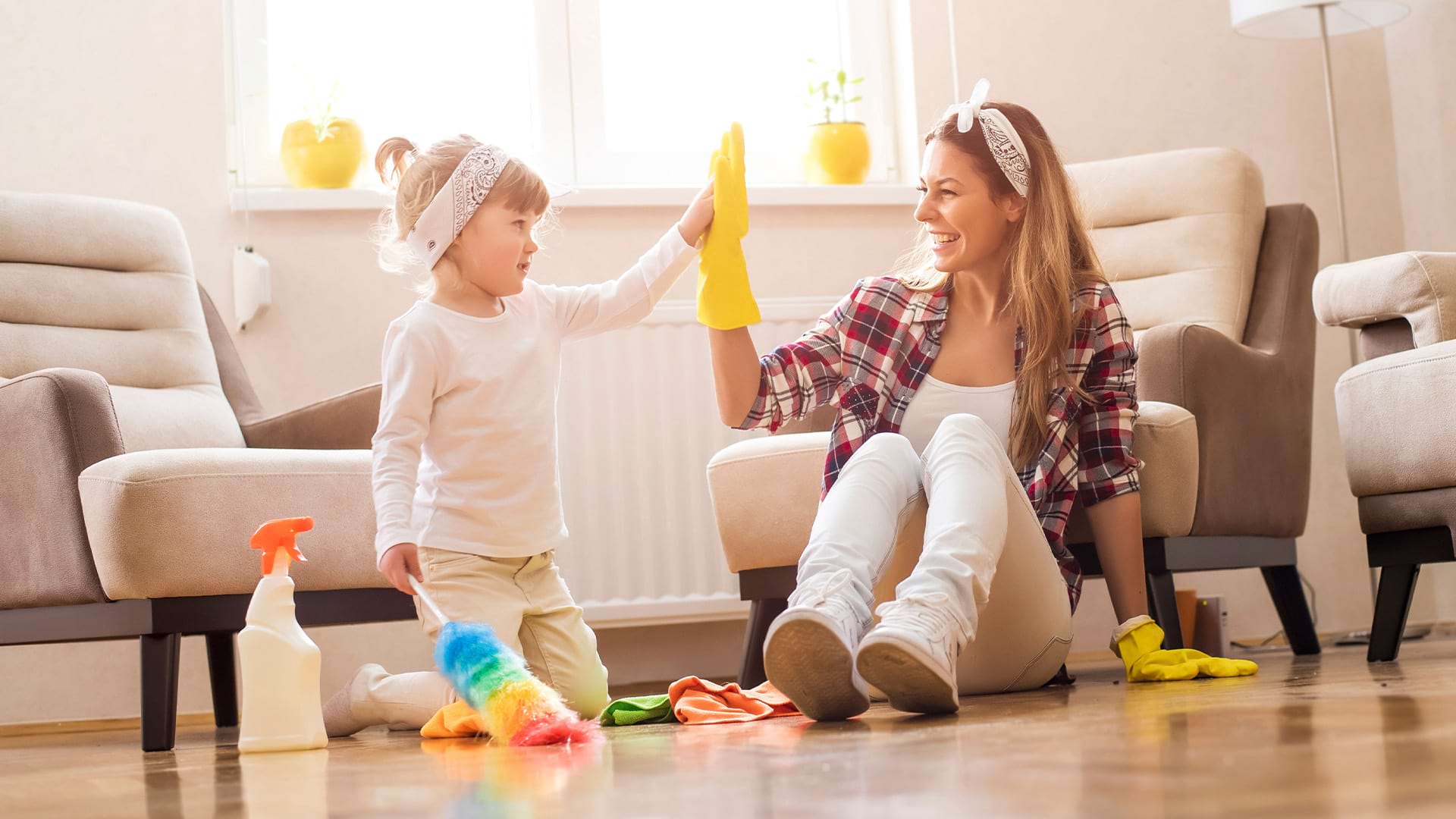 A smiling woman and a young girl sitting on a living room floor giving each other a high-five. The woman is wearing yellow rubber cleaning gloves, the girl is holding a colorful feather duster, and cleaning supplies are resting on the floor beside them.