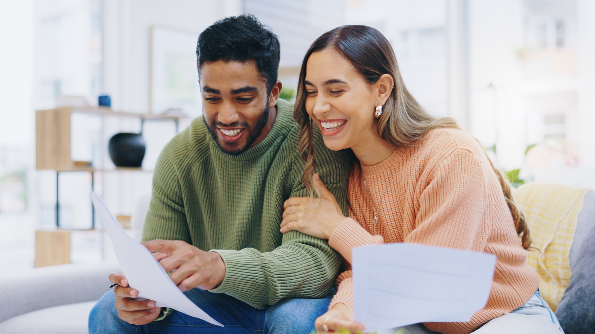 English A smiling couple sitting together on a couch, reviewing documents. The man is pointing at a paper he is holding with a pen, while the woman rests her hand affectionately on his arm and holds another document, both looking happy.