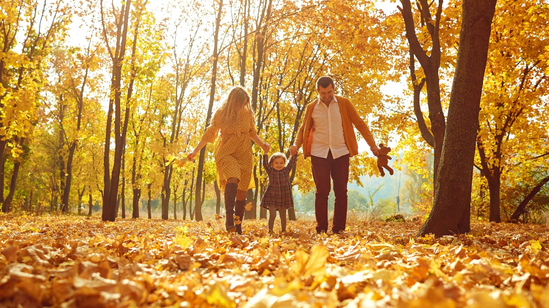 Family walking together along an outdoor path, with one adult carrying a baby in a child carrier and another adult holding a young child's hand.