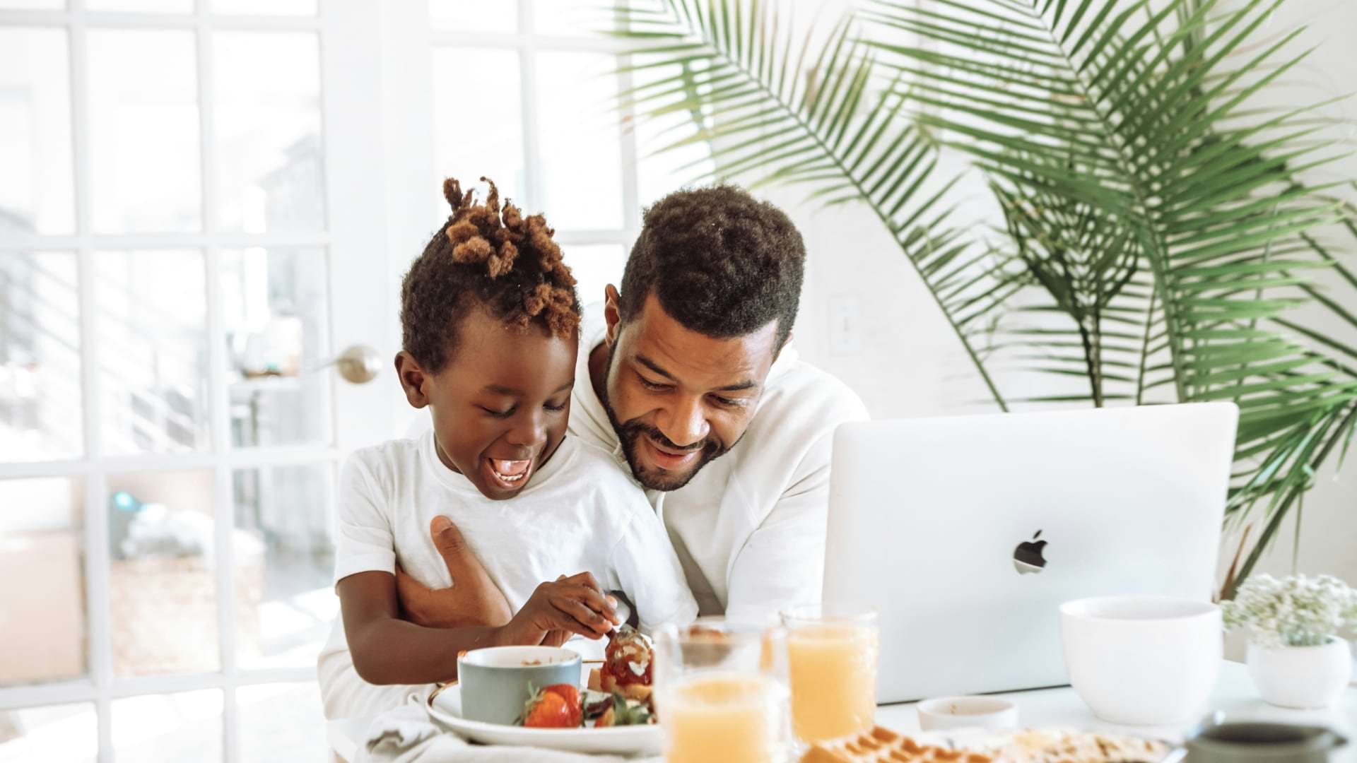 A child and an adult sit together at a breakfast table, sharing a meal and looking at a plate of fruit. A laptop, glasses of orange juice, and a houseplant are visible in the bright, sunlit room.