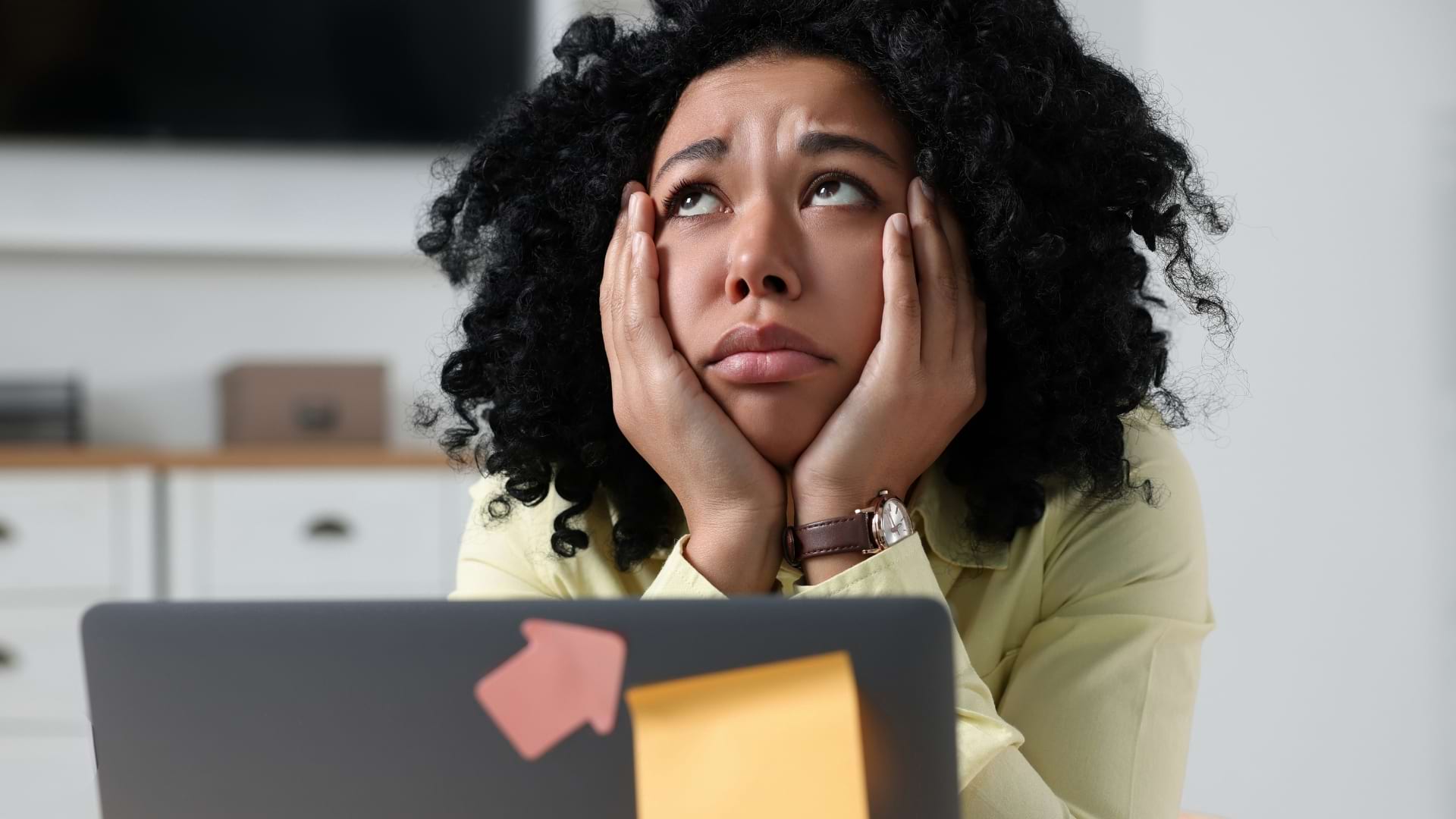 Person resting their face in their hands while sitting behind a laptop with sticky notes attached, suggesting concentration on administrative or household tasks.