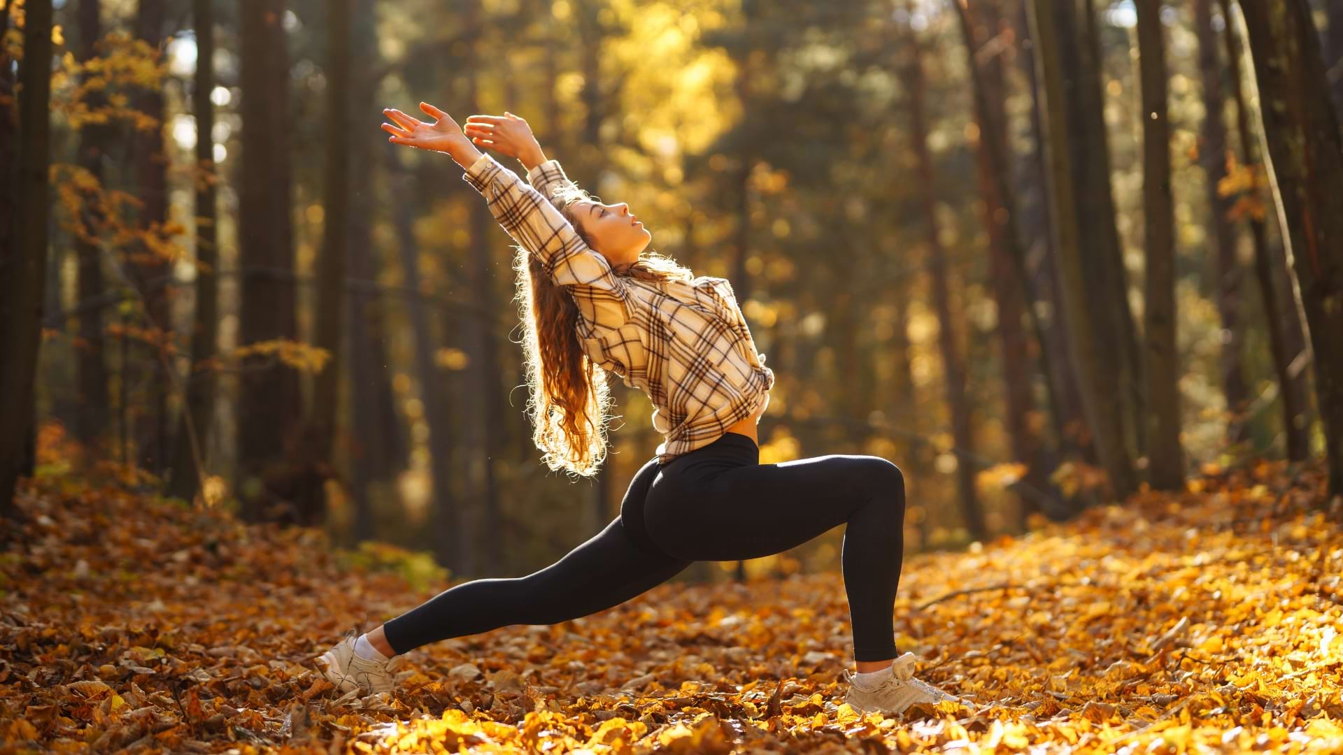A person performing a yoga lunge pose in a forest covered with autumn leaves, stretching both arms overhead. Warm golden sunlight filters through the trees, illuminating the woodland setting.
