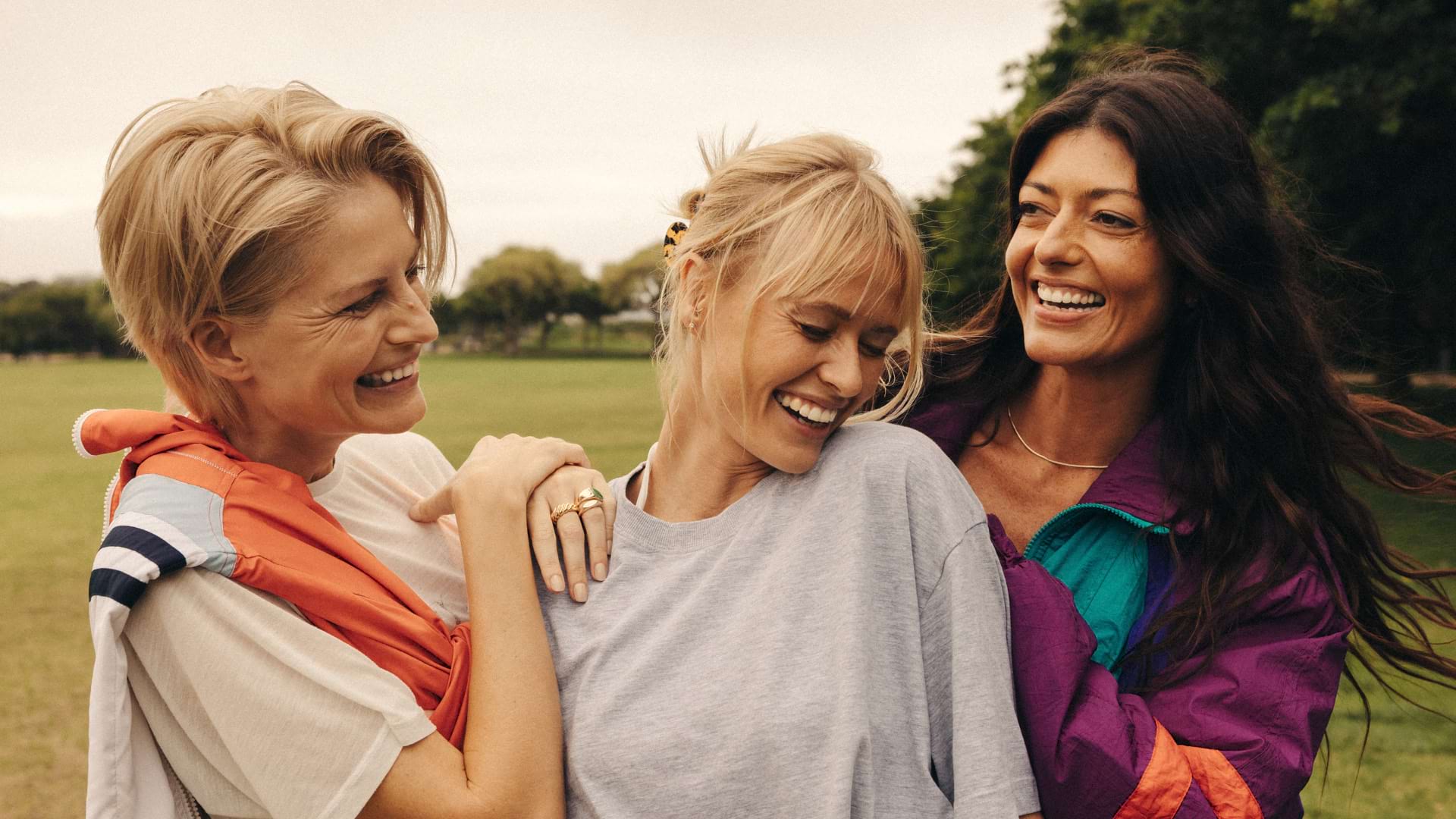Three friends stand close together outdoors on a grassy field with trees in the background. They’re smiling warmly at the camera. The person on the left has short blond hair and wears a light shirt with an orange jacket draped over their shoulders. The person in the middle has blond hair tied back and wears a light gray shirt. The person on the right has long dark hair and wears a colorful jacket with purple, teal, and orange accents.