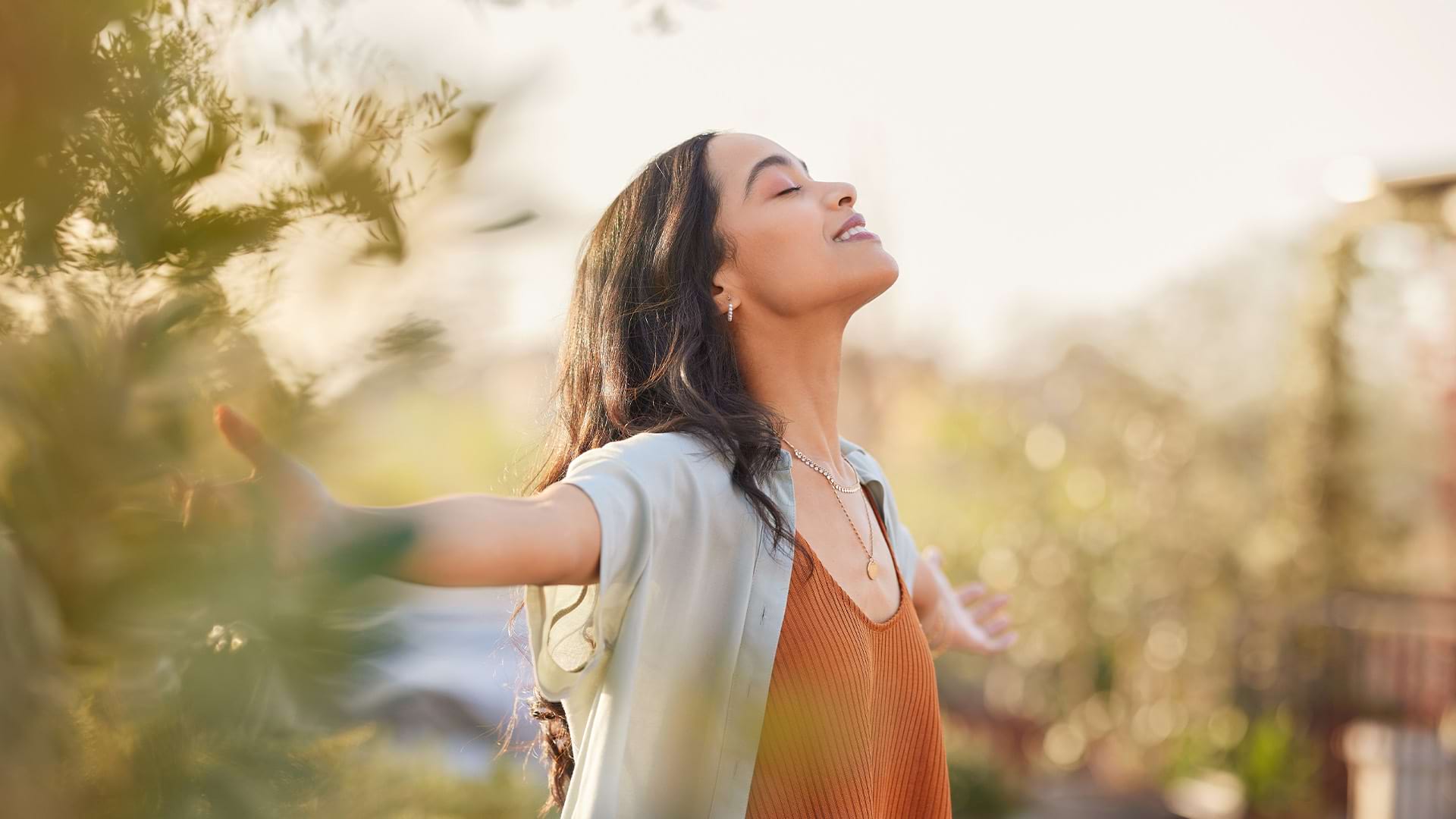 A young woman standing outdoors with her eyes closed and arms outstretched, taking a deep breath and enjoying the warm sunlight among green foliage.